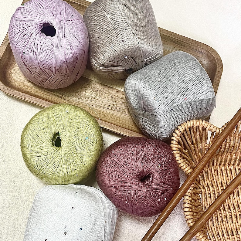 May include: Six balls of yarn in lavender, beige, silver, light green, dark pink, and white, each featuring small embedded sequins. The yarn balls are arranged on a wooden tray alongside a woven wicker basket and two long wooden knitting needles.