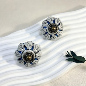 Peut inclure: Deux boutons de tiroir ronds en céramique avec une forme festonnée rappelant une fleur. La surface est ornée de motifs de pétales blancs et bleu profond, centrés par un élément métallique circulaire en laiton vieilli.