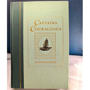May include: A hardcover book titled Captains Courageous by Rudyard Kipling with a light green cover, a dark green spine, and gold foil lettering. The front cover features a central illustration of a sailing ship inside an oval frame.