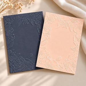 May include: Two rectangular cards, one dark navy blue and one light peach, each featuring an embossed floral border design. The raised patterns depict various flowers and leaves framing a smooth, blank central area on both paper surfaces.