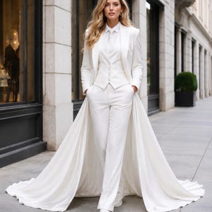 May include: A white three-piece suit comprising a tailored blazer, a button-up waistcoat, and straight-leg trousers. The blazer features a long, flowing detachable train that extends from the waist to the floor, creating a dramatic silhouette.