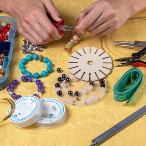 May include: A jewelry making kit featuring a round wooden kumihimo disk, various colorful gemstone beads, two spools of clear elastic cord labeled 1.0, a green measuring tape, metal pliers, and a plastic storage container filled with assorted beads.