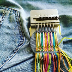 May include: A metal darning loom tool with a circular wooden base placed on blue denim fabric. The loom features vertical metal hooks holding multi-colored embroidery threads in shades of pink, orange, yellow, green, and blue, woven into a small square pattern.