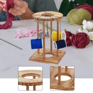 Op de afbeelding: Een houten garenkloshouder met een ronde bovenkant met ingekeepte randen en een vierkante voet. De standaard ondersteunt meerdere klossen gekleurd garen, waaronder blauw, geel en grijs, met draden die van de bovenkant naar de klossen lopen.
