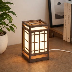 Könnte beinhalten: Eine rechteckige Tischleuchte mit einem dunkelbraunen Holzrahmen, der auf allen Seiten ein Gittermuster aufweist. Die Lampe hat eine weiße, durchscheinende Platte, die ein warmes, sanftes Licht ausstrahlt. Ein weißes Stromkabel führt aus dem Lampenfuß.