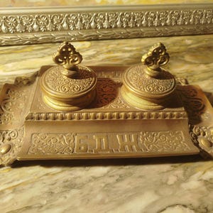 May include: A rectangular brass inkwell stand featuring two circular lidded ink containers with ornate filigree patterns. The base includes decorative scrollwork and the Cyrillic letters Б.Д.Ж. embossed on the front panel. The metal has a warm, aged gold-toned finish with intricate detailing throughout the entire structure.