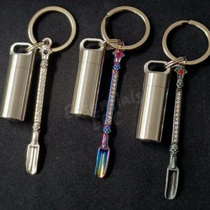 May include: Three metal keychains, each featuring a cylindrical container and a long, slender spoon attachment. The spoons are decorated with small skull motifs and embedded crystals, with one spoon in a rainbow finish and two in silver.