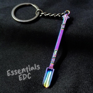 May include: A metal keyring featuring a small, iridescent, rainbow-coloured spoon with a decorative handle. The spoon has a polished finish with shifting hues of purple, blue, and gold. The text Essentials EDC is printed in white near the spoon.