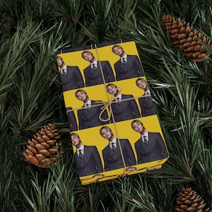 Puede incluir: Una caja de regalo rectangular envuelta en papel amarillo con un patrón repetitivo de una persona con traje gris oscuro de rayas diplomáticas, camisa blanca y corbata oscura. El paquete está atado con un cordel marrón fino sobre ramas de pino.