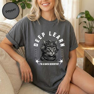 Puede incluir: Una camiseta gris carbón de manga corta con un gráfico blanco de un gato con equipo futurista. El texto dice DEEP LEARN e I'M A DATA SCIENTIST, con hebras decorativas similares al ADN flanqueando la ilustración del gato.