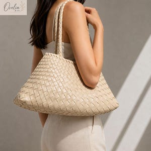 May include: A beige tote bag featuring a woven basket-weave pattern throughout the exterior. The bag includes two braided shoulder straps and a spacious, unstructured body. The design is consistent in color and texture, showcasing a uniform weave.