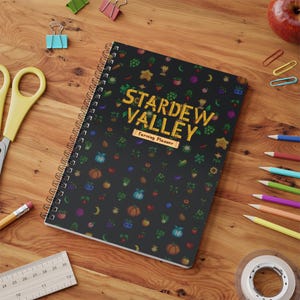 May include: A spiral-bound notebook with a black cover featuring a repeating pattern of colorful pixelated crops and the yellow text "STARDEW VALLEY" above a smaller rectangular label that reads "Farming Planner" in a dark brown font.