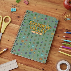 May include: A spiral-bound notebook with a sage green cover featuring a repeating pattern of pixelated crops and items from Stardew Valley. The center displays the text Stardew Valley Farming Planner in a yellow, pixelated font style.