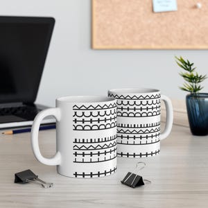 Puede incluir: Dos tazas de cerámica blanca con un patrón geométrico negro repetitivo que consiste en bandas horizontales de triángulos, semicírculos y líneas entrecruzadas. Las tazas tienen un cuerpo cilíndrico con un asa curva en el lateral.