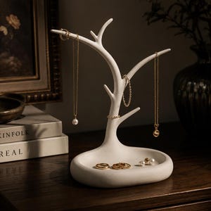 May include: A matte white jewellery stand shaped like a tree with multiple branches for hanging necklaces and bracelets. The base is a smooth, oval-shaped tray designed to hold rings and earrings. Several gold-toned necklaces and rings are displayed.