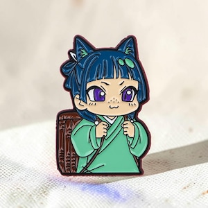 Può includere: Una spilla in smalto metallico raffigurante un personaggio in stile chibi con capelli blu scuro, orecchie da gatto e occhi viola. Il personaggio indossa una veste verde chiaro e porta un cesto intrecciato marrone sulla schiena.