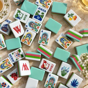May include: A collection of rectangular Mahjong tiles featuring a light green back and white face. The tiles display colorful illustrations of buildings, flowers, plants, and the word JOKER, along with numbers and letters like BAM, W, and CRAK.
