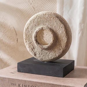 May include: A circular, beige, porous stone sculpture with a central hole, mounted on a solid, rectangular black base. The object features a rough, textured surface throughout, contrasting with the smooth, flat finish of the dark pedestal.