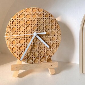 May include: A circular wall clock featuring a light brown woven cane webbing face and three simple white hands. The clock is supported by a small, light-coloured wooden tripod easel stand, allowing it to be displayed upright on a flat surface.