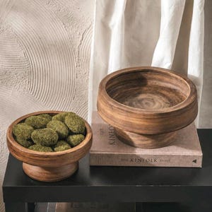 Könnte beinhalten: Zwei runde Holzschalen auf einem Sockel mit dunkelbraunem Finish und sichtbarer Holzmaserung. Eine Schale ist mit mehreren kleinen, texturierten grünen Mooskugeln gefüllt. Die Schalen stehen auf einem hellbraunen Buch namens The Kinfolk Table.