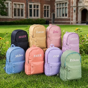 Peut inclure: Une collection de huit sacs à dos de différentes couleurs unies, notamment noir, beige, rose, lavande, bleu clair, corail, violet et vert sauge. Chaque sac à dos possède une poche avant zippée et un prénom brodé en blanc ou en rose.