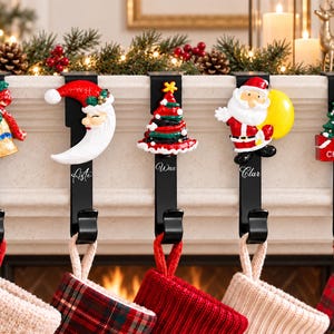 May include: Five black metal stocking holders with decorative Christmas icons including a gold bell, a crescent moon face, a striped tree, Santa Claus, and a green tree with the text Merry Christmas. Each hook holds a hanging holiday stocking.