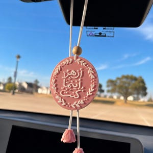 May include: A round pink car hanging ornament featuring white Arabic calligraphy text surrounded by a white leaf border. The ornament is suspended by white cords with a small wooden bead and two pink flower-shaped tassels at the bottom.