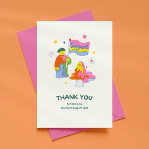May include: A white greeting card featuring a colorful cartoon illustration of a person holding a flag that says Support, next to a melting blob character. The text below reads THANK YOU for being my emotional support idiot in dark green.