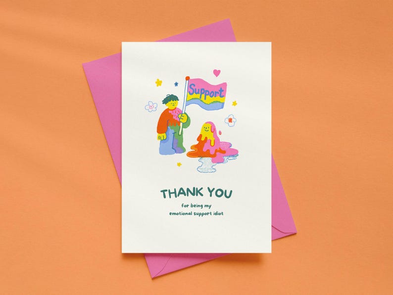 May include: A white greeting card featuring a colorful cartoon illustration of a person holding a flag that says Support, next to a melting blob character. The text below reads THANK YOU for being my emotional support idiot in dark green.