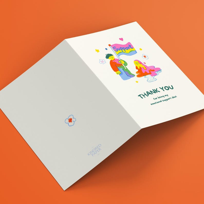 May include: A folded white greeting card featuring a colorful illustration of a figure holding a flag labeled Support, next to a melting blob character. The text reads THANK YOU For being my emotional support idiot. A small flower icon is on the back.