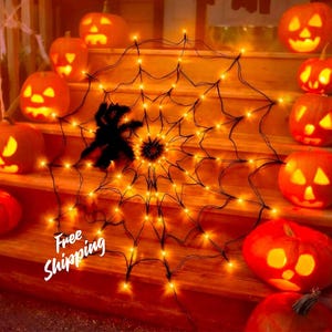May include: A circular black spider web decoration illuminated with warm orange LED lights, featuring a fuzzy black spider at the centre. The web is surrounded by several glowing orange jack-o'-lanterns with carved faces on wooden steps.