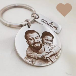 Peut inclure: Un porte-clés en métal argenté avec un pendentif circulaire gravé d'un portrait photographique d'un adulte et d'un enfant. Une petite étiquette rectangulaire attachée à l'anneau est gravée du mot custom dans une police d'écriture cursive.