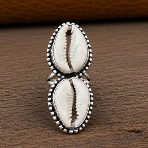 Puede incluir: Un anillo de plata con dos conchas de cauri blancas apiladas verticalmente. Cada concha está engastada en un marco de plata con cuentas, creando un diseño alargado. El metal tiene un acabado pulido que resalta la textura natural.