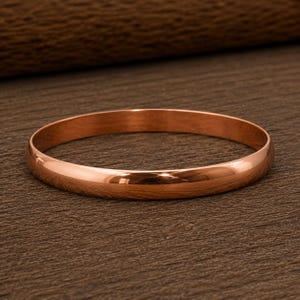 May include: A polished copper bangle bracelet with a smooth, rounded surface and a continuous circular design. The metal has a warm, reddish-brown metallic luster that reflects light evenly across its curved exterior, creating a simple, solid band.