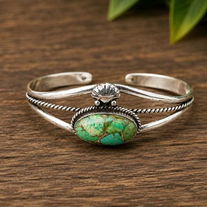 May include: A silver cuff bracelet featuring a central oval-shaped green and brown marbled stone. The band is constructed from two smooth silver wires and one twisted rope-style wire, accented with a small silver shell motif above the stone.