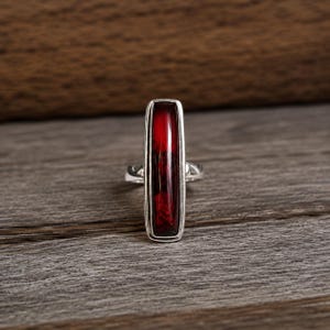 May include: A silver ring featuring a long, rectangular, deep red stone set vertically. The metal band is smooth and polished, framing the elongated, translucent red gemstone which displays subtle internal variations in color and texture.