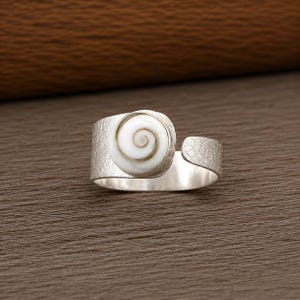 May include: An adjustable silver ring featuring a textured band and a circular white shell centrepiece with a natural brown spiral pattern. The metal surface has a matte finish, and the ring is open at the back to allow for size adjustment.