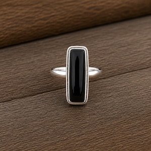 May include: A silver ring featuring a long, vertical, rectangular black onyx stone set in a smooth metal bezel. The band is polished silver, and the stone has a flat, reflective surface that contrasts with the metallic frame of the ring setting.
