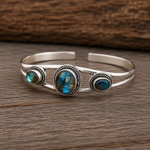 May include: A silver cuff bracelet featuring three oval-shaped labradorite gemstones with blue and gold iridescence. The stones are set in twisted rope-style silver bezels, and the band consists of two parallel silver wires that meet at the ends.