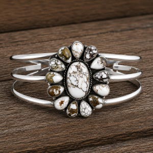 May include: A silver cuff bracelet featuring a central oval stone surrounded by twelve smaller teardrop-shaped stones. The stones are white with brown and black matrix patterns. The bracelet has a triple-band design made of polished silver metal.