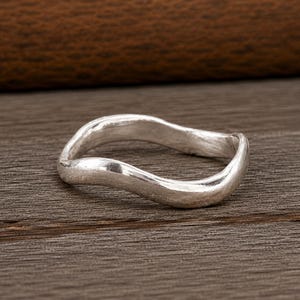 以下が含まれることがあります： 連続した波状のバンドデザインが特徴のシルバーリング。金属の表面はマットでわずかに質感のある仕上げになっており、リングの曲線に沿って柔らかな光沢を生み出しています。バンドは全体を通して一定の厚みを保っています。
