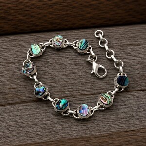 Könnte beinhalten: Ein silberfarbenes Kettenarmband mit acht ovalen Abalone-Muschel-Einlagen in schillernden Grün-, Blau- und Violetttönen. Das Armband verfügt über einen Karabinerverschluss und eine kurze Verlängerungskette mit runden Gliedern zur Größenanpassung.