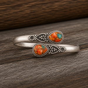 May include: A bypass cuff bracelet crafted from oxidized silver, featuring two teardrop-shaped settings at each end. Each setting contains a vibrant orange and turquoise stone inlay, accented by intricate metal scrollwork and beaded detailing.