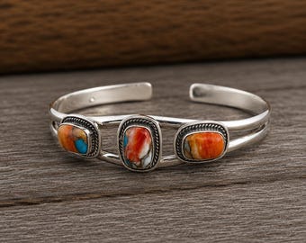 May include: A silver cuff bracelet featuring three oval-shaped stones with vibrant orange, white, and blue marbled patterns. Each stone is set in a polished silver bezel with a twisted rope border, connected by a smooth, open-ended silver band.