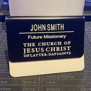 May include: A rectangular black name badge with white text reading JOHN SMITH, Future Missionary, and THE CHURCH OF JESUS CHRIST OF LATTER-DAY SAINTS. The badge features a white bottom section and is placed against a dark computer keyboard.