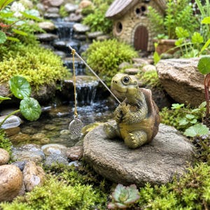 May include: A small, green, textured turtle figurine sitting on a rock, holding a thin metal fishing rod with a gold-colored chain and a silver-toned fish charm dangling from the end of the line, designed for garden or outdoor display.