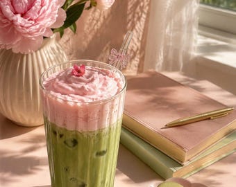 May include: A tall, ribbed glass filled with iced green matcha topped with a thick layer of pink foam and a single small flower. A clear glass straw with a pink bow detail sits in the drink. Beside it are two green and one pink macaron.