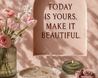 May include: A fluted glass vase with pink ranunculus and tulips, a beige organic-shaped stone tray holding gold jewelry, a green ribbed candle, and a pink silk scrunchie. An arched wall niche features the text: TODAY IS YOURS. MAKE IT BEAUTIFUL.