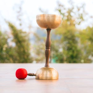 Pode incluir: Uma taça tibetana de latão montada sobre uma base de pedestal de madeira castanho-escura, fixada a um suporte de latão em forma de cúpula. Um martelo de madeira com uma ponta de feltro vermelho vivo repousa na superfície junto à base.