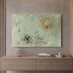 May include: A rectangular wall art piece featuring a pale sage green background with abstract markings in black, brown, and blue. The composition includes thin, scribbled lines and scattered paint splatters across the surface, creating a layered effect.